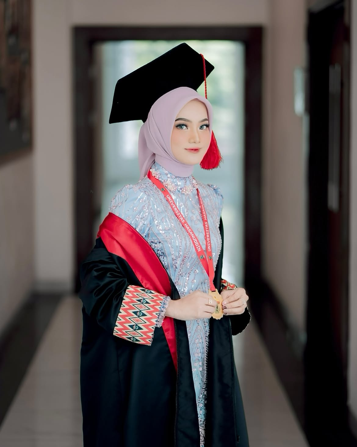 Rias wisuda dengan toga hitam, selempang merah, dan hijab lavender